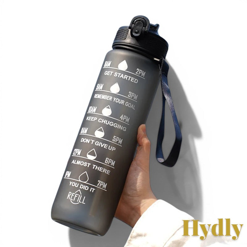 Gr8est 1L black motivational water bottle with time markers and straw held against a blue sky