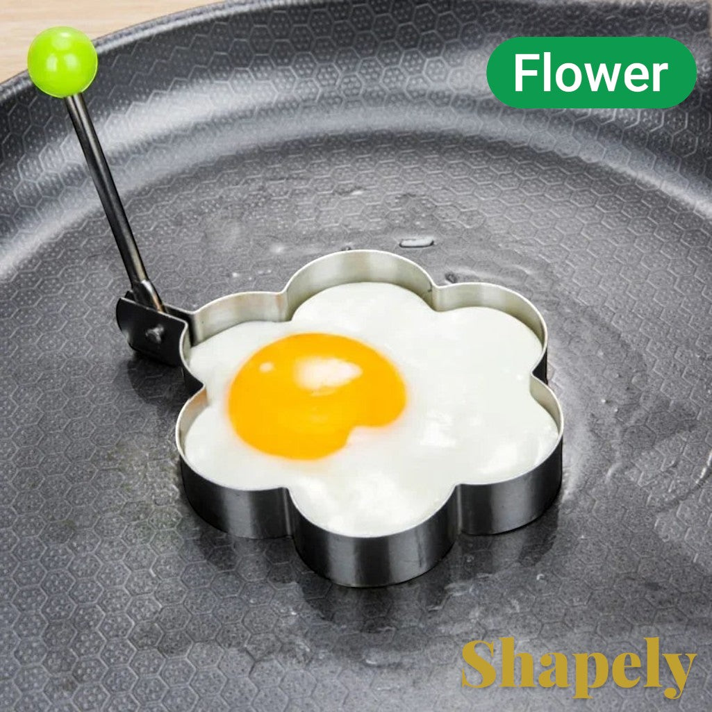 Gr8est stainless steel flower-shaped egg mold cooking ring with a green handle on a non-stick frying pan.