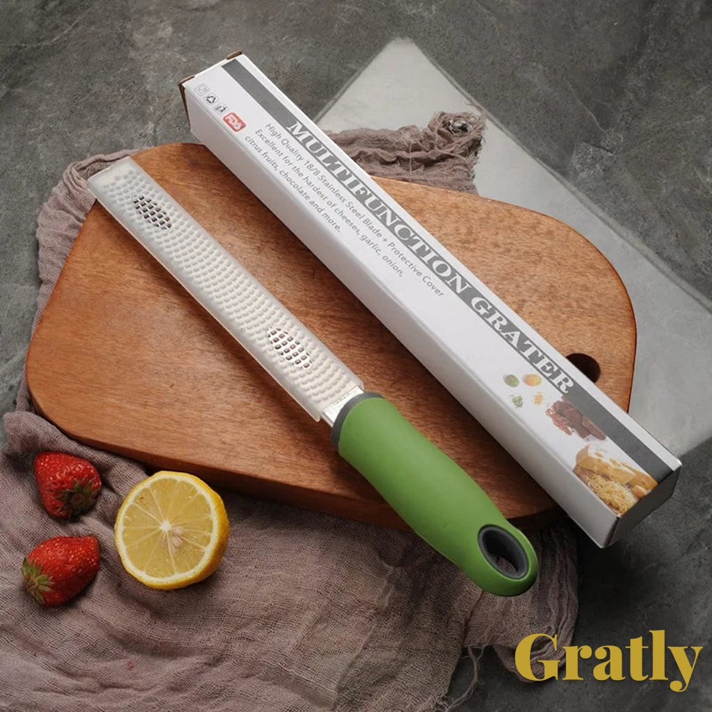 Gr8est Gratly stainless steel multi-purpose grater with a green ergonomic handle, shown with its box on a wooden board.
