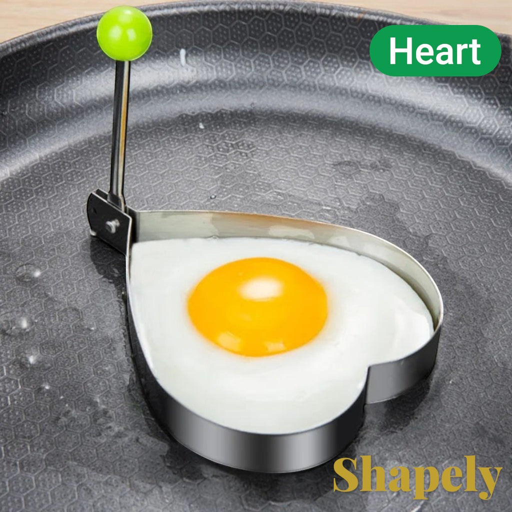 Gr8est heart-shaped stainless steel egg mold with a green handle cooking a fried egg in a non-stick pan.