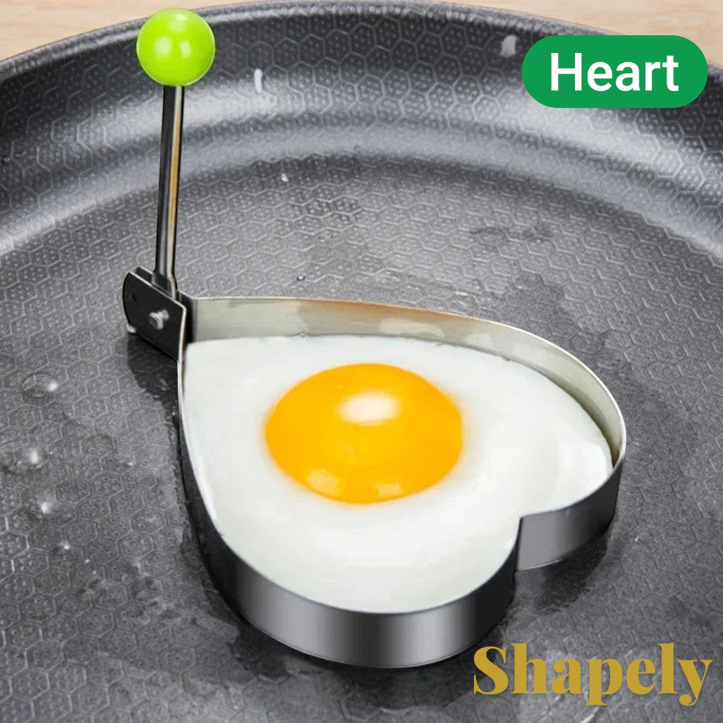 Gr8est stainless steel heart-shaped egg mold with a green handle on a non-stick frying pan