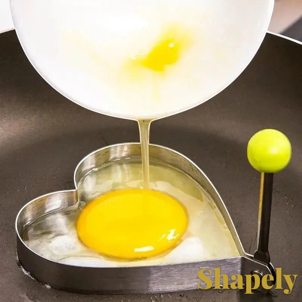 Gr8est heart-shaped stainless steel egg mold with a green handle being used to cook an egg in a frying pan.