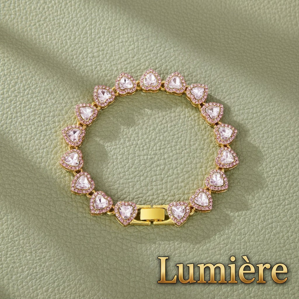 Gr8est Lumière heart tennis bracelet in gold with pink rhinestone accents on a textured green background.