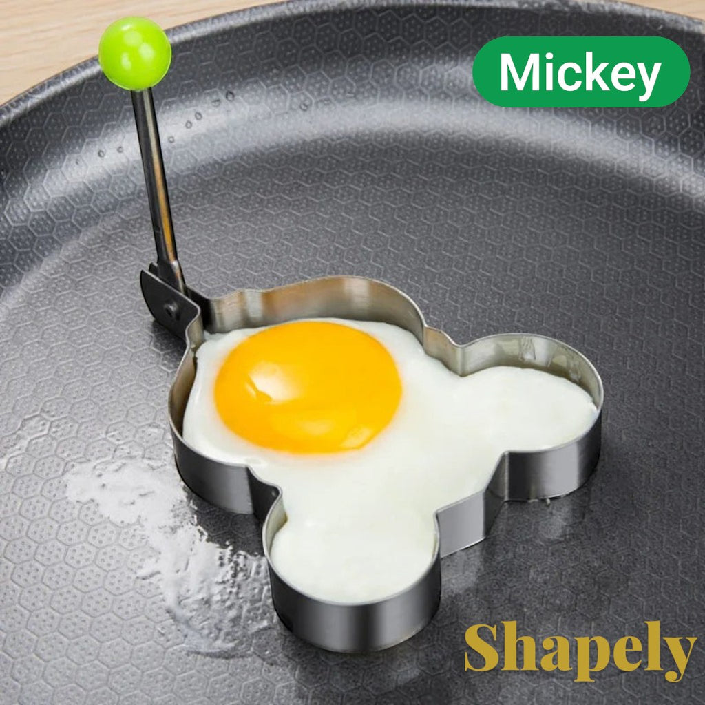 Gr8est stainless steel Mickey Mouse shaped egg mold with a green handle on a non-stick frying pan.