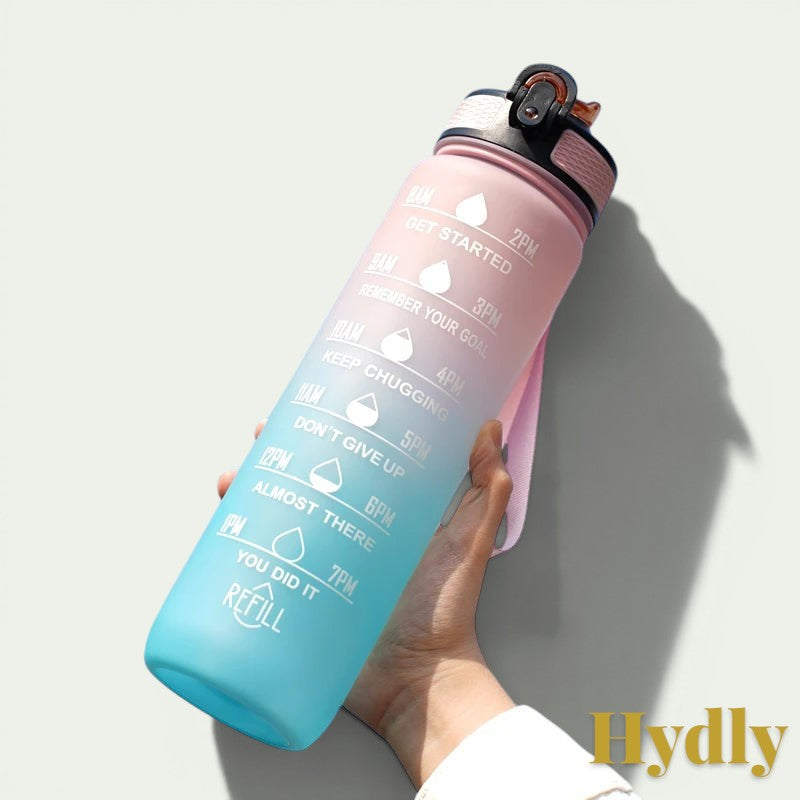 Gr8est 1L motivational water bottle in pink and blue gradient with time markers and straw held against blue sky