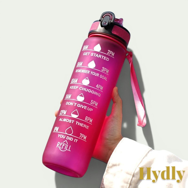 Gr8est 1L pink motivational water bottle with time markers, straw, and leak-proof lid held against a blue sky.