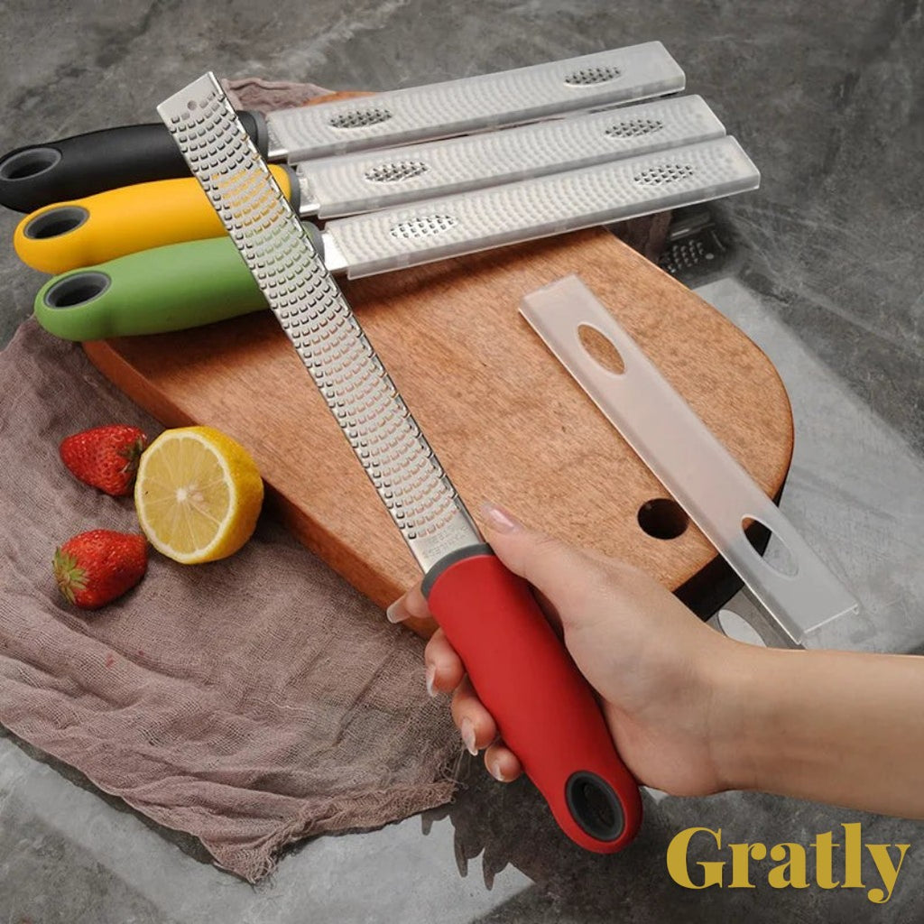 Gr8est Gratly stainless steel multi-purpose kitchen grater with a red ergonomic handle held over a wooden board.