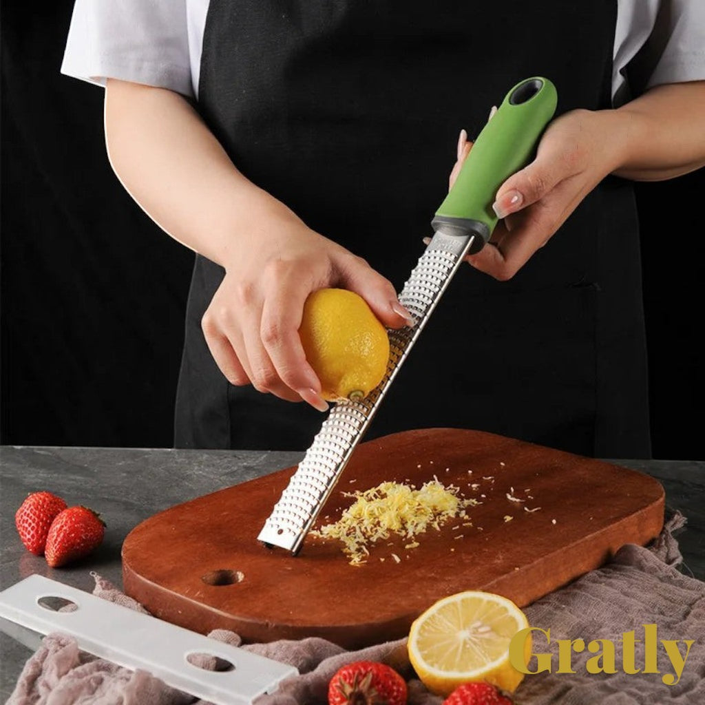 Gr8est stainless steel multi-purpose grater with green ergonomic handle zesting a fresh lemon on a wooden board